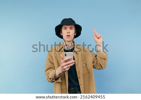 Surprised young man in panama and shirt stands on blue background with smartphone in hand and shows finger up with shocked face looking at camera.