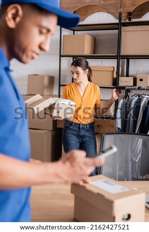 Young seller holding carton box near clothes and blurred african american deliveryman in online web store