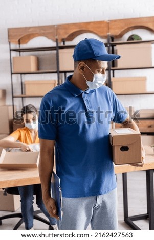 African american courier in medical mask holding cardboard box near blurred owner of online web store