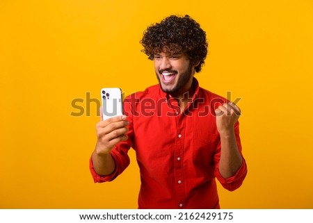 Happy satisfied man with beard holding smartphone and smiling making yes gesture, celebrating online lottery or giveaway victory. Indoor studio shot isolated on orange background Royalty-Free Stock Photo #2162429175