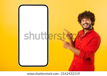 Playful handsome curly indian bearded man points fingers at huge smartphone with empty blank screen isolated on yellow, happy latin guy presenting mobile app, showing best deal, copy space, template Royalty-Free Stock Photo #2162432119