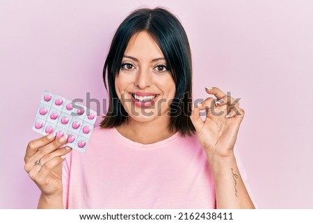 Young hispanic woman holding pills doing ok sign with fingers, smiling friendly gesturing excellent symbol 