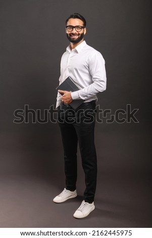 Full length of happy Indian bearded man in smart casual wear and stylish glasses standing isolated on black background and carrying digital tablet Royalty-Free Stock Photo #2162445975