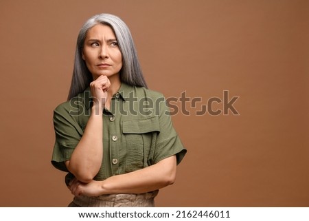 Smart and thoughtful mature woman holding her chin and pondering idea, making difficult decision, looking uncertain doubtful. Indoor studio shot isolated on beige background Royalty-Free Stock Photo #2162446011