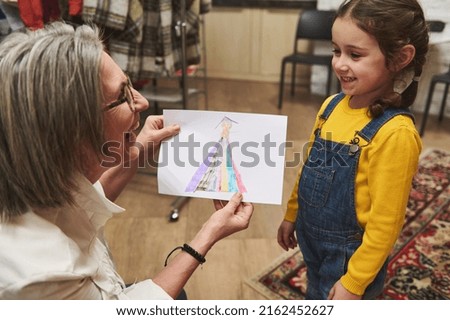 Pleasant elderly Caucasian woman tailor, caring and loving grandmother smiles admiring the clothes sketch of her adorable granddaughter while working in a fashion design studio and tailoring atelier