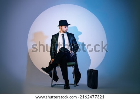 Handsome private detective sitting on chair in the spotlight on studio background. noir film style. Spy, investigation concept. 