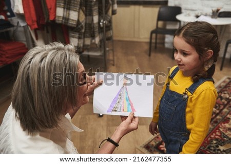 Focus on an adorable baby girl. Loving grandmother   smiles to her charming granddaughter and admires her beautiful drawing - a sketch of clothes. Fashion design concept, art, creativity, hobby
