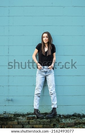 A teenage girl with long hair wearing black, denim and combat boots leaning against a blue cinder block wall and looking away with attitude.