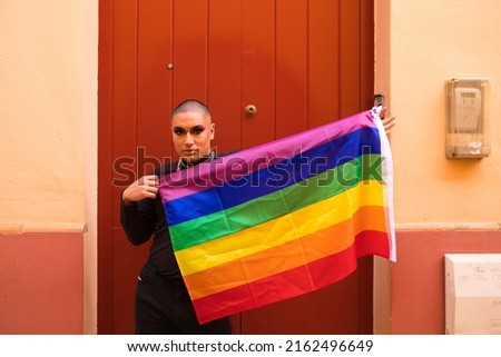 Non-binary person, of South American origin and young, the person is wearing make-up and holding the gay pride flag on a red background. Concept equality, homosexuality, gay, lesbian, gay pride.