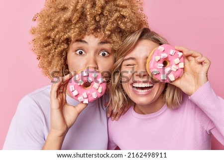 Photo of two female best friends hold delicious doughnuts with marshmallow have sweet tooth enjoy eating tasty unhealthy food dressed casually isolated over pink background. Harmful nutrition Royalty-Free Stock Photo #2162498911
