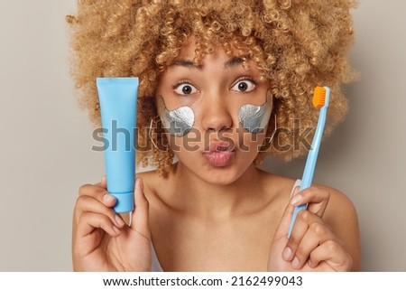 Headshot of surprised curly haired woman keeps lips folded widely opened eyes holds toothpaste and toothbrush cleans teeth regularly undergoes hygiene procedures isolated over grey background