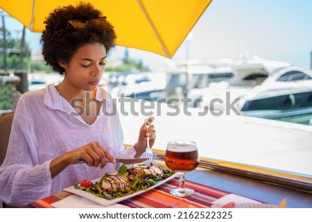 Young black female, with afro hair, enjoying eating a goat cheese salad in a bar in a coastal area, with boats in the background.