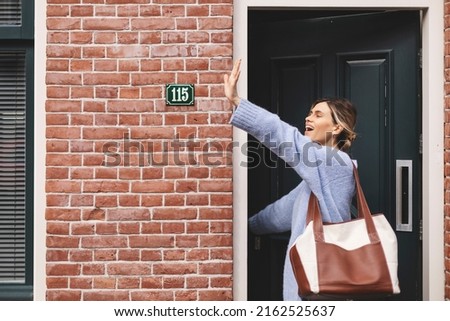 Smiling young woman waving with a friendly cheerful smile to her new neighbours. Girl leaves the house closing the door and waving her hand. Girl wear blue sweater and brown bag, meeting friends. Royalty-Free Stock Photo #2162525637