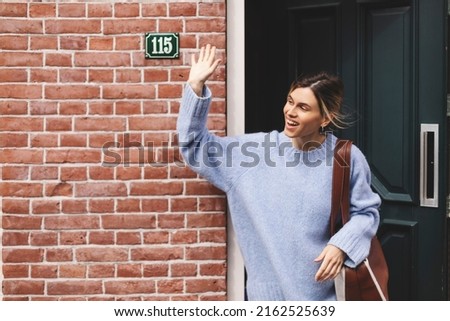 Smiling young woman waving with a friendly cheerful smile to her new neighbours. Girl leaves the house closing the door and waving her hand. Girl wear blue sweater and brown bag, meeting friends. Royalty-Free Stock Photo #2162525639