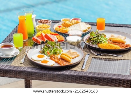 Hotel breakfast. Pair having breakfast on tropical villa near swimming pool. Table full of food - exotic fruits, eggs and bakery at luxury hotel in hot summer day. Balanced food Royalty-Free Stock Photo #2162543163