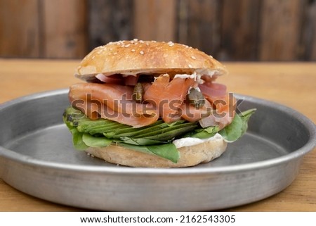 Gastronomy. Closeup view of a smoked salmon sandwich made with bagel bread, pink salmon filet, cream cheese, sliced avocado, capers, onion and arugula, in a metal dish on the wooden table.