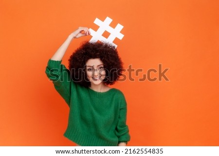 Portrait of funny positive woman with Afro hairstyle wearing green casual style sweater standing holding hashtag above her head, smiling. Indoor studio shot isolated on orange background.