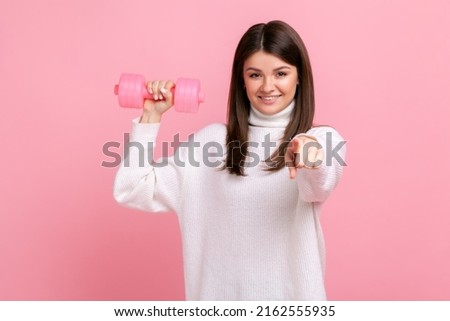 Smiling female holding rose dumbbells and pointing finger to camera, calls on to go in for sport, wearing white casual style sweater. Indoor studio shot isolated on pink background.