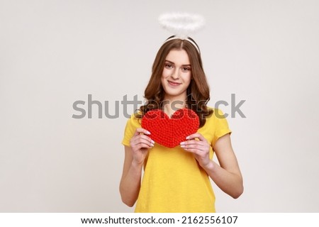 Portrait of adorable charming teenager girl in yellow T-shirt with angelic nimbus holding red paper heart and looking smiling at camera. Indoor studio shot isolated on gray background.