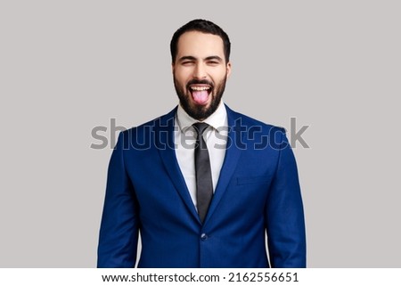 Portrait of playful bearded businessman showing tongue, having fun, fooling around, childish manners, wearing official style suit. Indoor studio shot isolated on gray background.