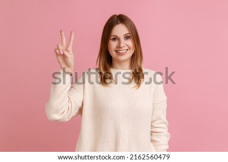 Woman showing victory or peace gesture, smiling broadly, rejoicing lucky winning, feeling optimistic about future success, wearing white sweater. Indoor studio shot isolated on pink background.