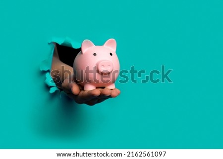 hand holds a piggy bank blue background out of a torn hole in a paper background 