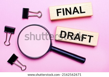On a pink background, a magnifier, black paper clips and wooden blocks with the text FINAL DRAFT. Business concept