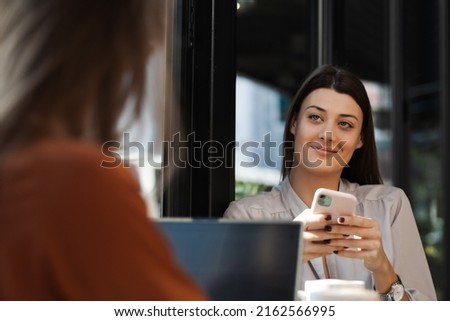 Two young business women in a cafe having one on one meeting. Friends after work talking gossiping and having coffee at a window table on a sunny day.