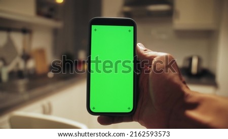 Man holding mobile phone with green screen at night in kitchen. Mock up chroma key surfing internet, watching content video.