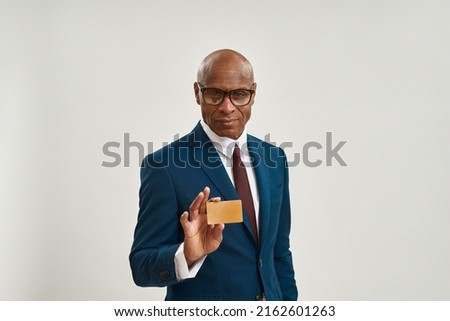 Serious black businessman holding and showing golden business card. Adult man wearing formal wear and glasses. Modern successful male lifestyle. Isolated on white background in studio. Copy space