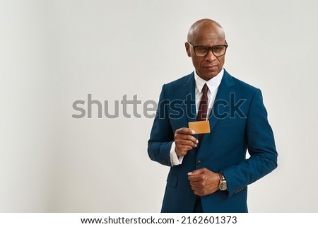 Serious black businessman showing golden business card. Bald adult man wearing formal wear and glasses looking at camera. Modern successful male lifestyle. White background in studio. Copy space