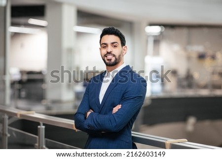 Smiling confident calm attractive young middle eastern businessman with beard in suit with crossed arms looks at camera in office interior. Successful business, ceo manager and leadership at factory Royalty-Free Stock Photo #2162603149