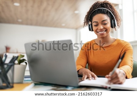 Smiling young black woman in wireless headphones sitting at desk table working on laptop and writing letter in paper notebook, taking notes watching weninar, holding pen in hand, free copy space Royalty-Free Stock Photo #2162603317