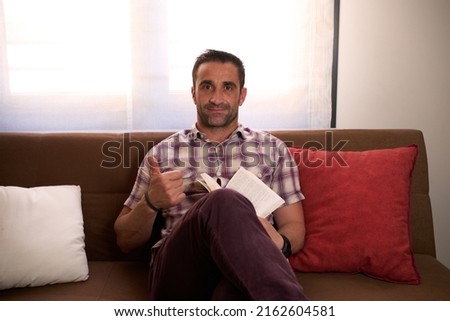 Man in a plaid shirt and book in his hand sitting relaxed on a sofa