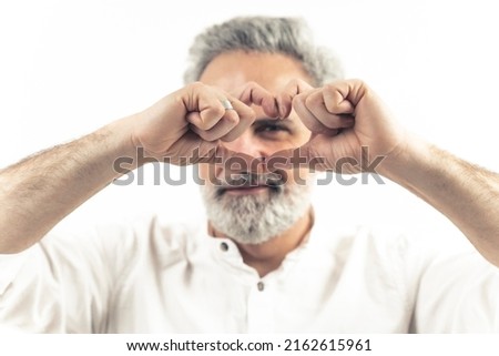 Studio shot on white background of an european attractive man in his 60s making a heart sign with his fingers over his face. Focus on the foreground. High quality photo