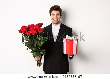 Concept of holidays, relationship and celebration. Image of handsome smiling guy in black suit, holding bouquet of red roses and giving present on new year, white background