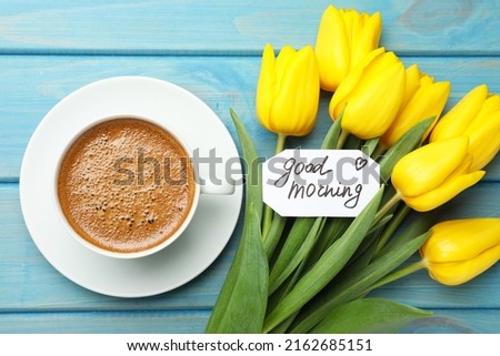 Cup of aromatic coffee, beautiful yellow tulips and Good Morning note on light blue wooden table, flat lay