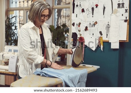 Positive elderly gray-haired Caucasian woman ironing shirt, working in a fashion design and repair atelier. Fashion designer, seamstress, dressmaker, tailor workplace. Pinned sketches on studio wall