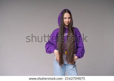 Nice cute young girl with long hair in bright hood posing on gray background. Charming girl in a lilac sweatshirt smiles and poses on isolated.