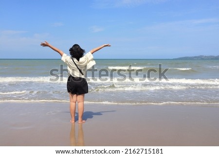 Back view of Asian short black hair woman raise hands with the sea, wave beat, colorful sky and white clouds background. Nature, freedom, happiness, powerful woman concept.