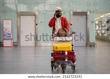 Young smiling African American man pushing luggage trolley while walking after arrival at airport, talking on mobile phone. Happy Black male tourist rolling a baggage cart in terminal. Trip, journey. Royalty-Free Stock Photo #2162723241