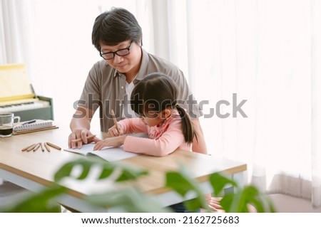 Portrait of Asian father and cute little daughter sitting at table drawing a picture or writing book with colour pencils at home. Happy to spend time togetherness at home. Hobby concept.