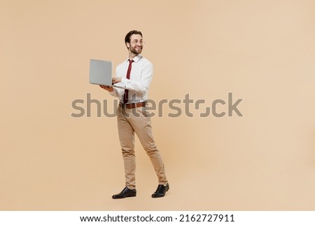 Full body young employee business man corporate lawyer 20s wear white shirt red tie glasses work in office hold use laptop pc computer look aside on worspace area isolated on plain beige background