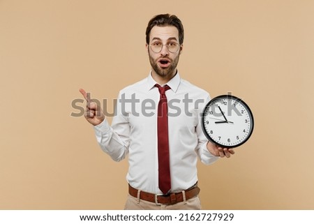 Young shocked employee business man corporate lawyer 20s wearing white shirt red tie glasses work in office hold clock point finger aside on workspace area isolated on plain beige background studio