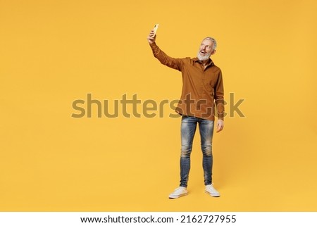 Full size body length excited smiling fancy elderly gray-haired bearded man 60s years old wears brown shirt doing selfie shot on mobile cell phone isolated on plain yellow background studio portrait