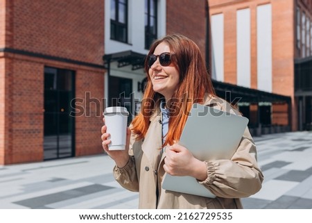 Caucasian redhead businesswoman in fashion office clothes posing outside with a laptop and eco cup of tea. Mockup, Woman using of the notebook computer and holding with bio white coffee cup
