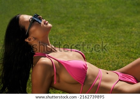 Side view of seductive female lying on lawn, sunbathing. Pretty brunette girl wearing pink swimsuit and sunglasses, enjoying vacation, looking up. Concept of summertime and youth.