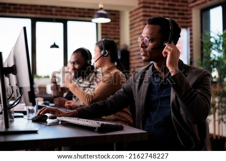 Male helpline assistant working at call center reception with multiple monitors at helpdesk. Male operator using telecommunication to help clients at customer service support, remote network. Royalty-Free Stock Photo #2162748227