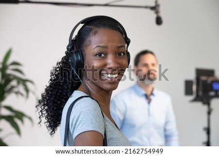 African American Woman Smiling Wearing Headphones Working as an Audio Person on a Video Production Set. Interview Subject, Microphone and Lighting in Background