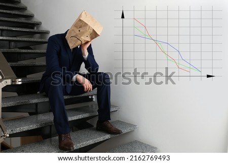 Man wearing paper bag with drawn sad face indoors and illustration of falling down chart. Economy recession concept